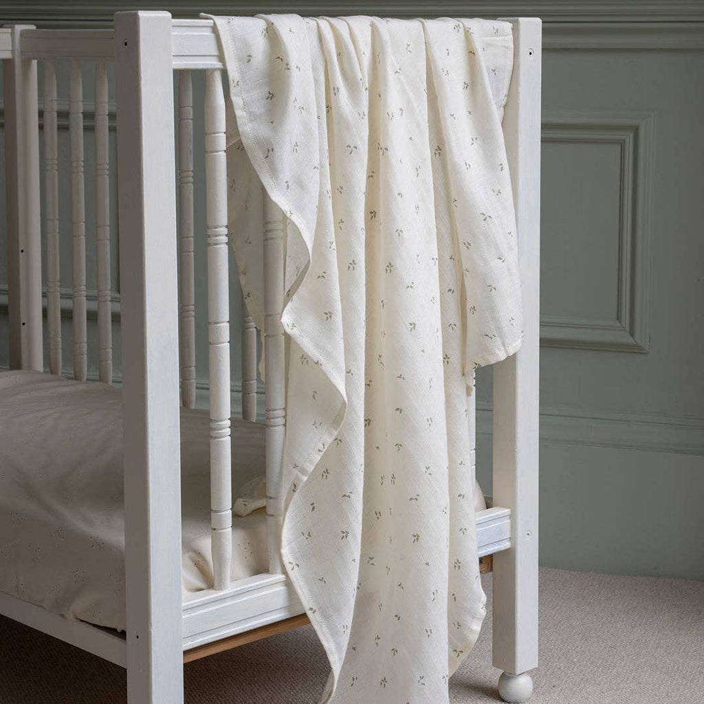 White crib with a Avery Row Swaddle muslin draped over the side against a gray wall.