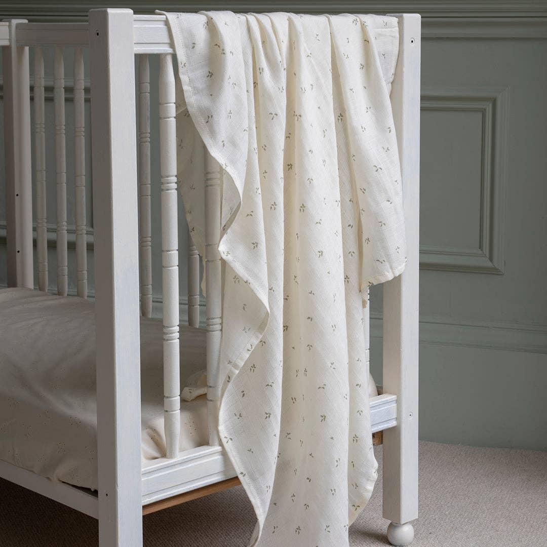 White crib with a Avery Row Swaddle muslin draped over the side against a gray wall.