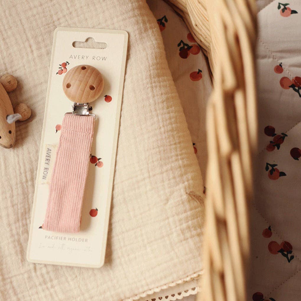 Baby pacifier holder with pink holder on Avery Row packaging against a soft fabric background