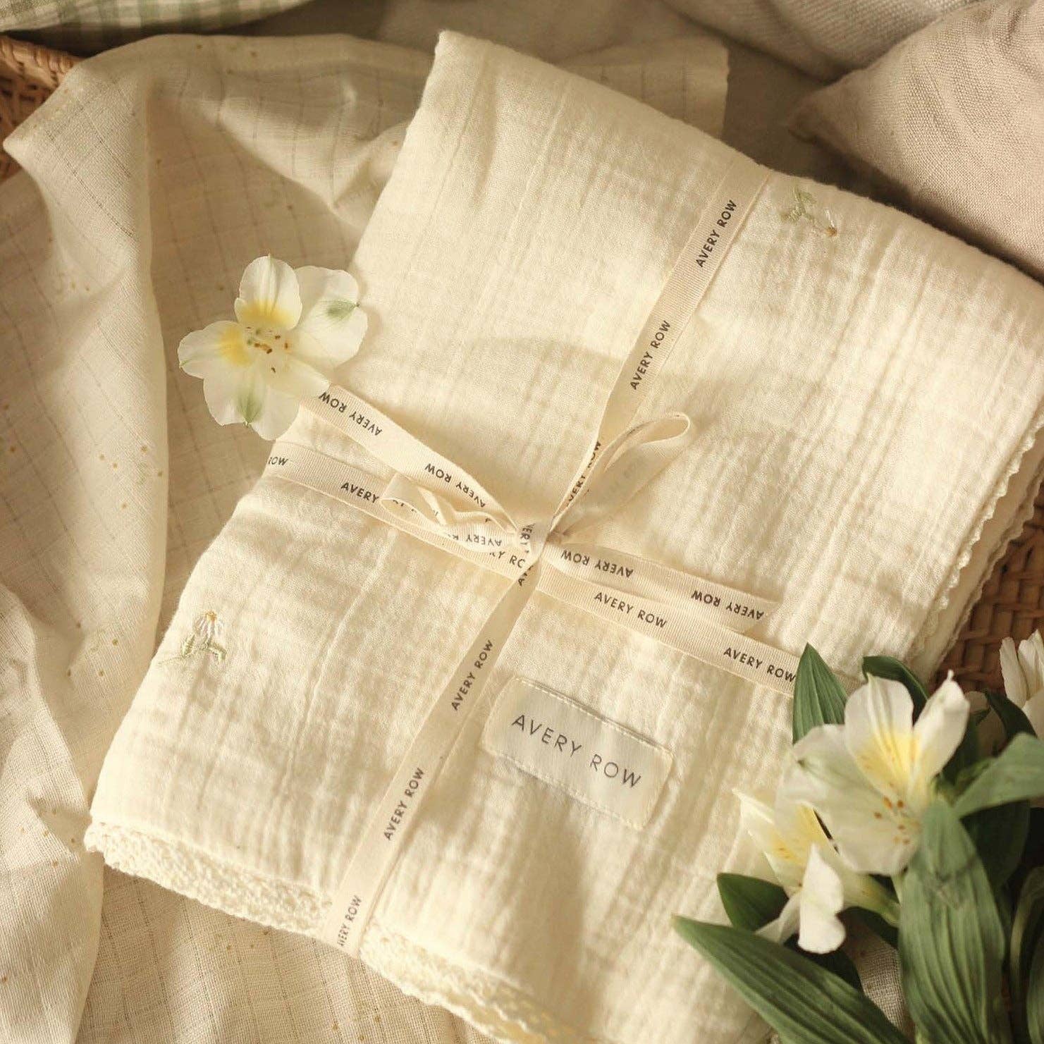 Folded muslin blanket with 'Avery Row' branding, tied with a ribbon, surrounded by flowers on a textured surface.