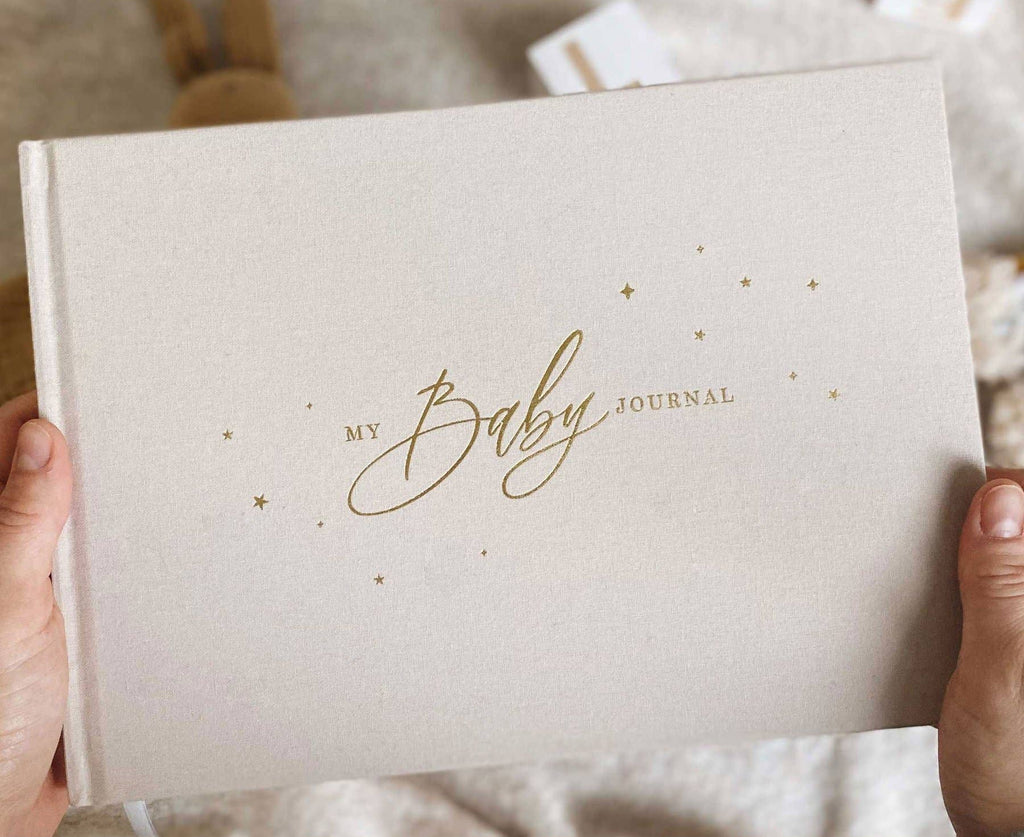 Beige journal titled 'My Baby Journal' held by hands on a soft fabric background.