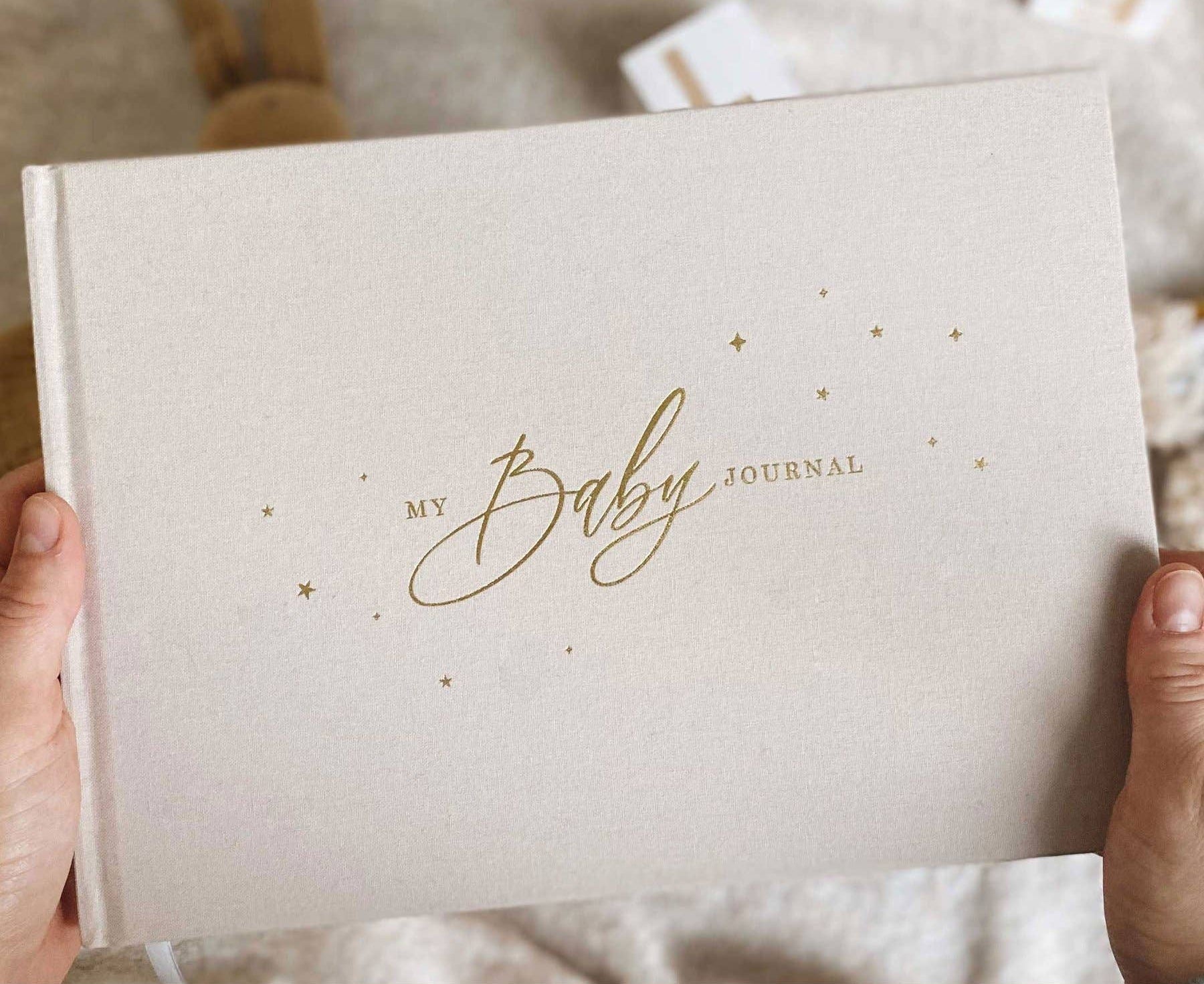 Beige journal titled 'My Baby Journal' held by hands on a soft fabric background.
