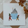 Card with illustration of two characters pushing a large blue cupcake with a candle, on a textured surface with brown envelope and dried flowers.