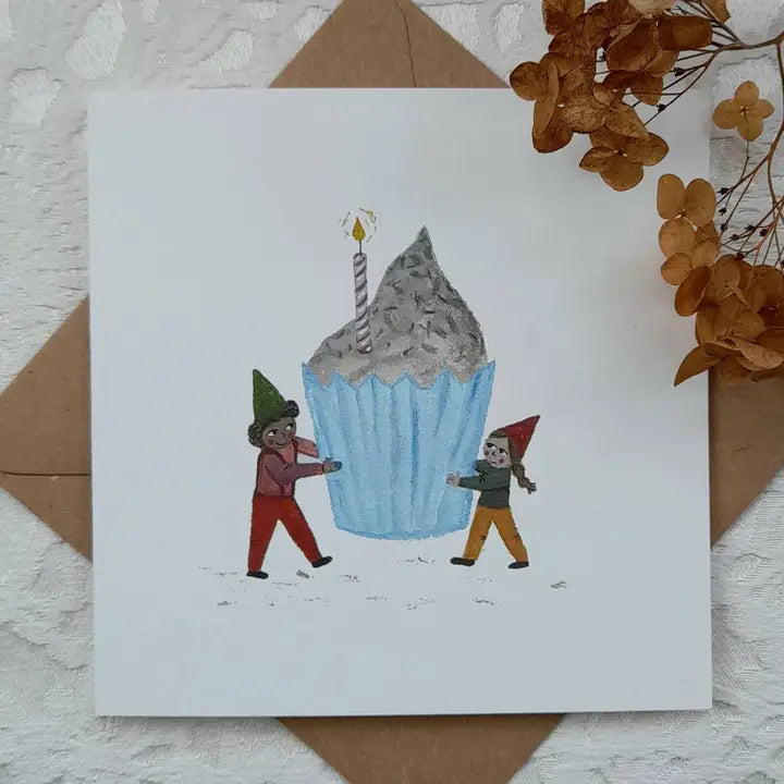 Card with illustration of two characters pushing a large blue cupcake with a candle, on a textured surface with brown envelope and dried flowers.