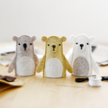 Three small bear figurines on a wooden surface with crafting materials.