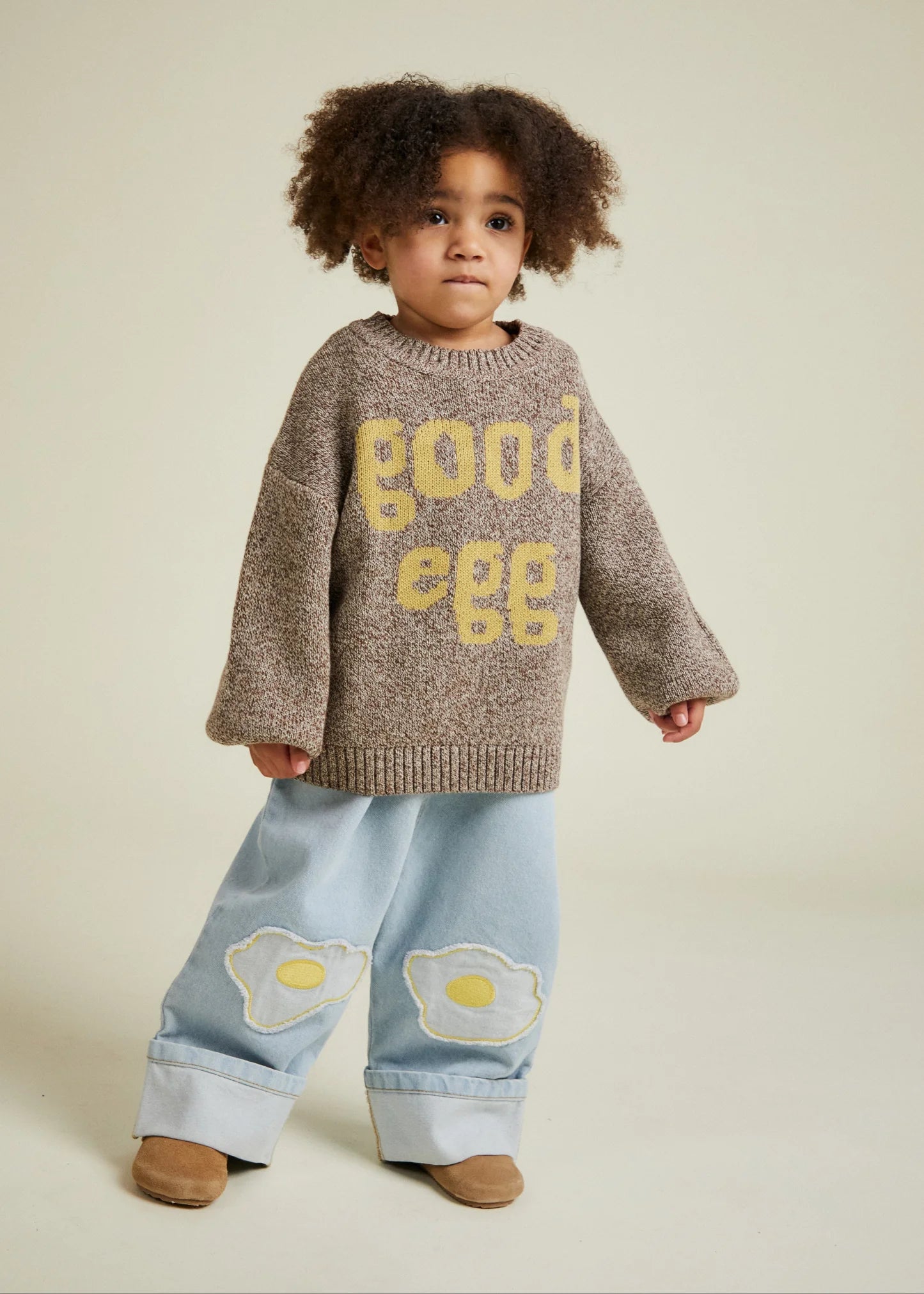 Child wearing a brown sweater with yellow text and light blue pants with egg designs on a beige background