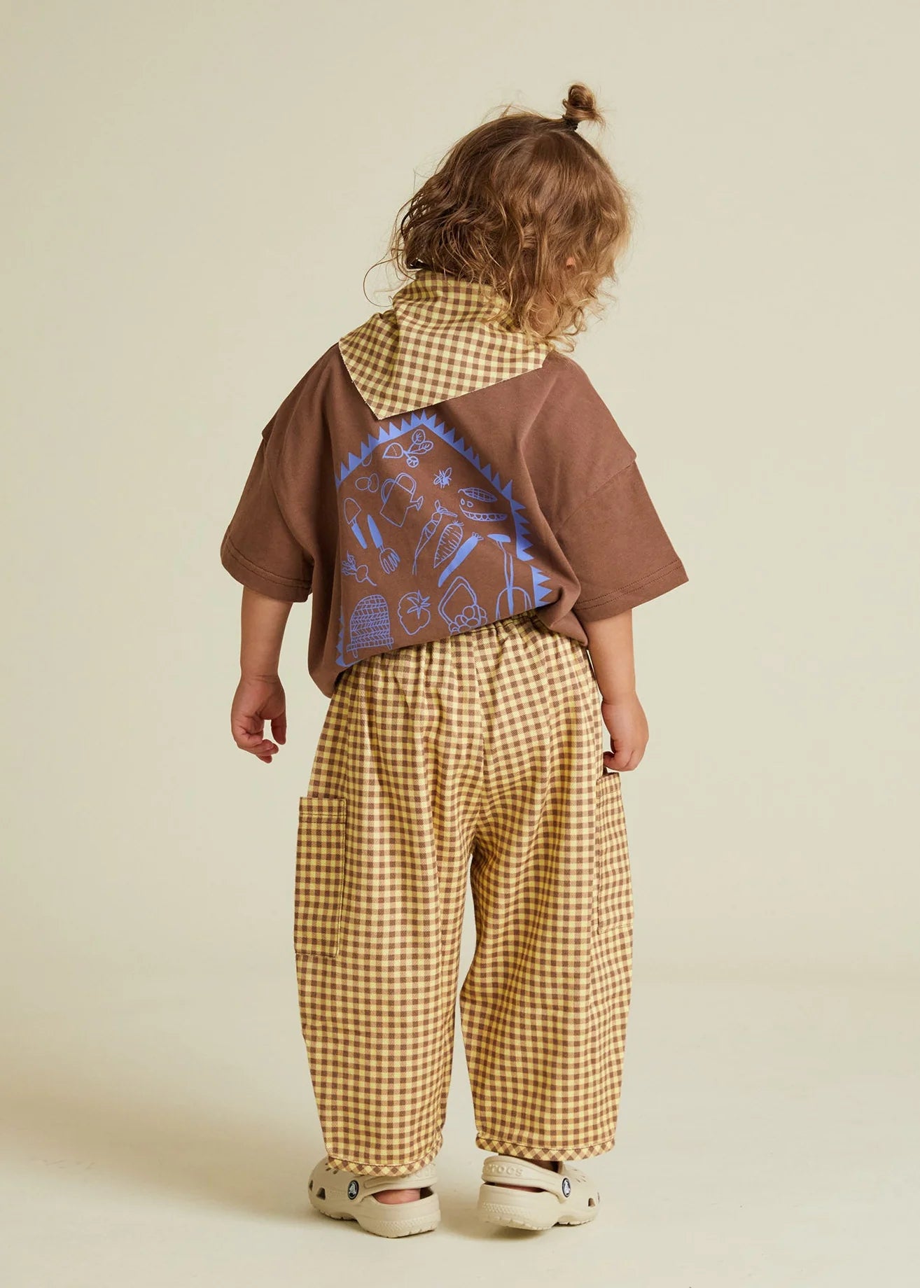 Child wearing a brown shirt with a colorful back design and gingham trousers on a beige background