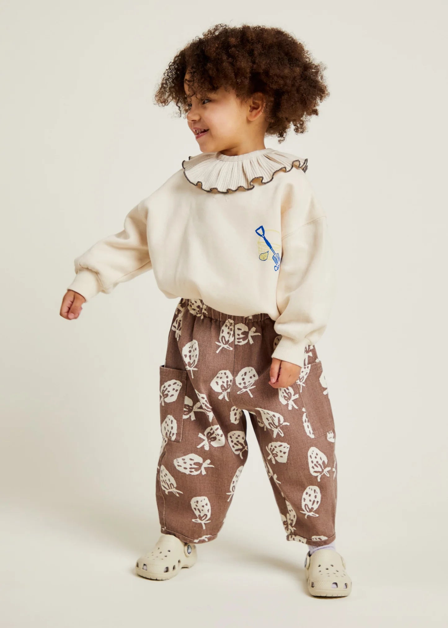 Child wearing a cream sweater with a ruffled collar and brown strawberry patterned pants on a white background