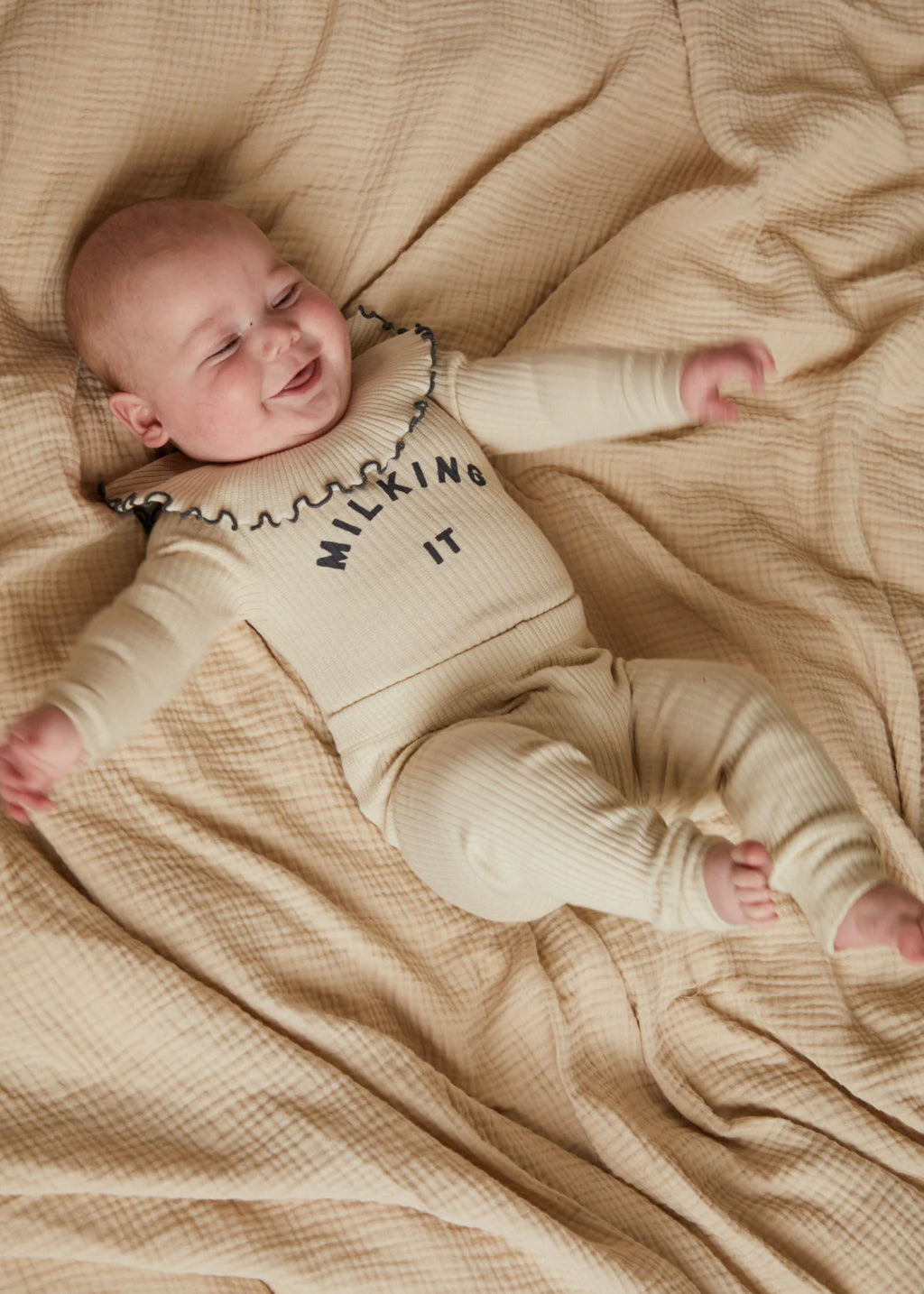 Baby lying on a beige blanket wearing a matching outfit with text.