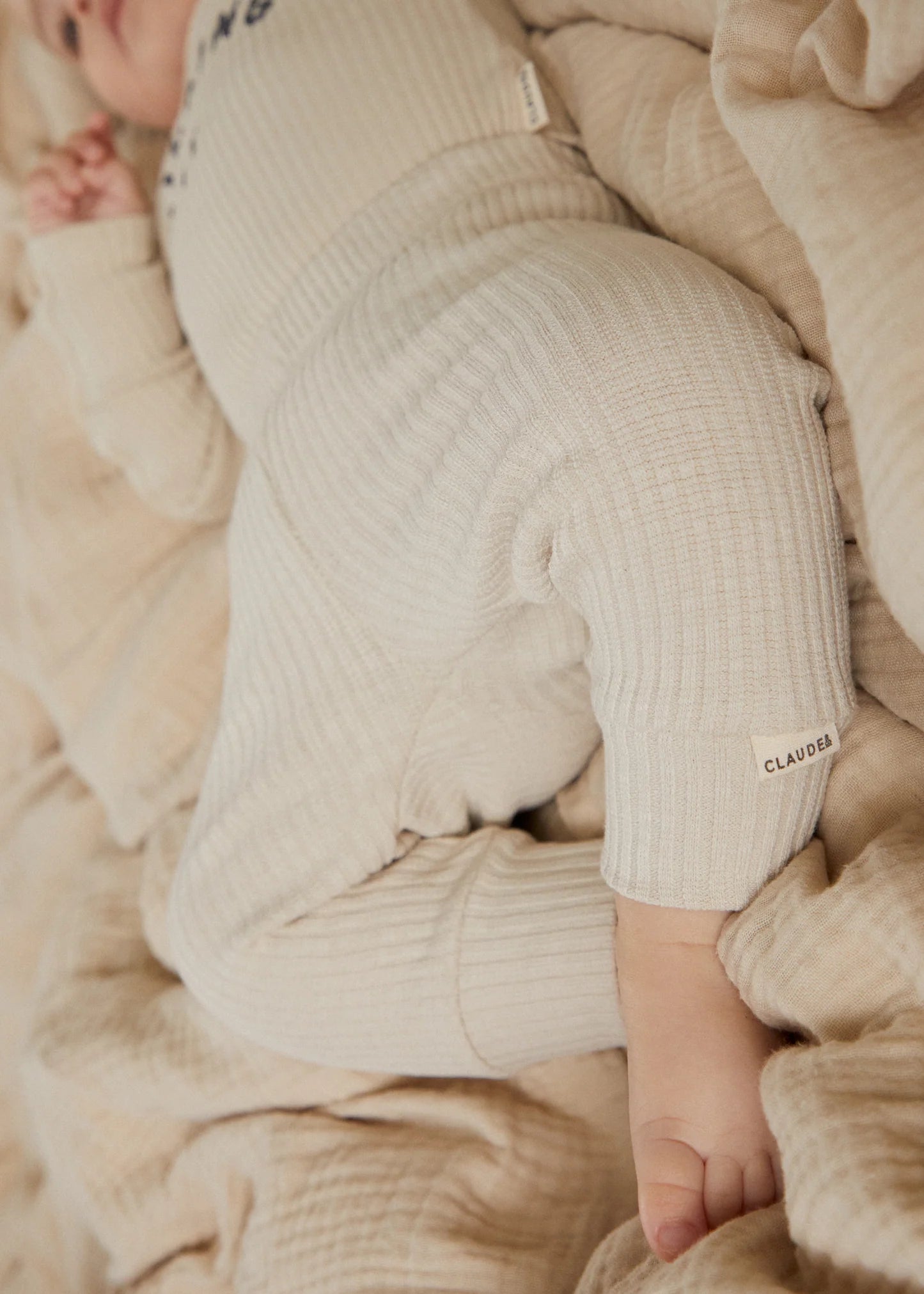 Close-up of a baby in a beige ribbed outfit lying on a beige blanket.
