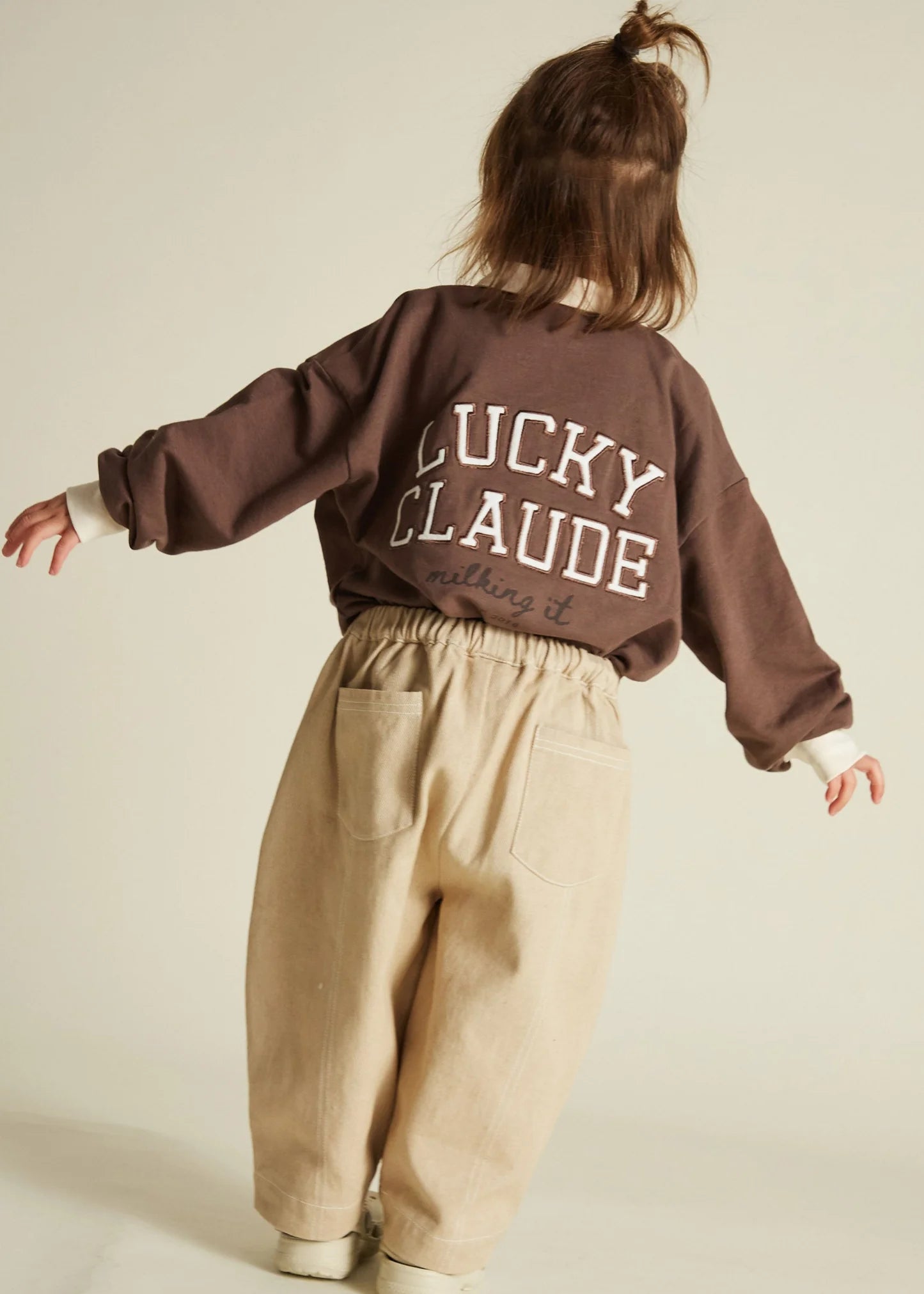 Child wearing a brown sweatshirt with 'Lucky Claude' text and sand trousers on a beige background