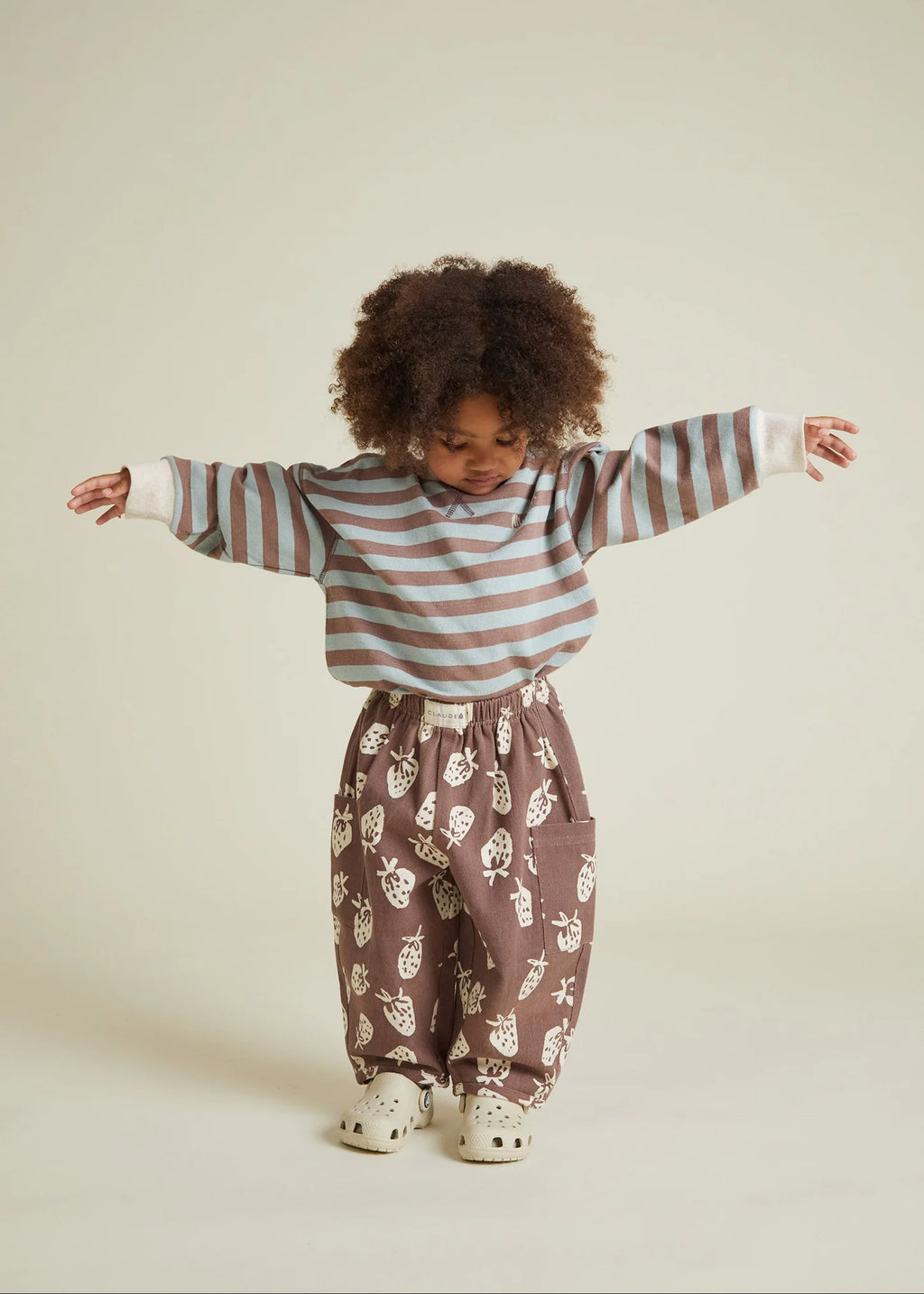 Child wearing a striped sweater and strawberry patterned pants on a plain background