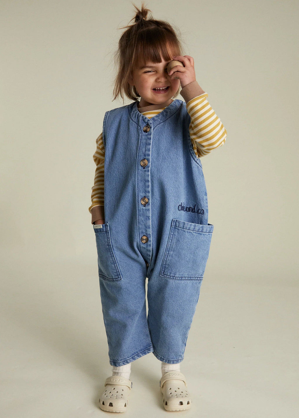 Child wearing a denim overall with a striped shirt underneath on a plain background