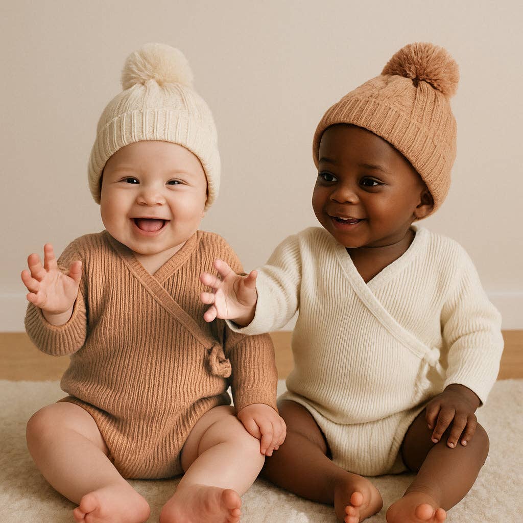 Two babies wearing matching knit hats and rompers sitting on a light-colored floor.