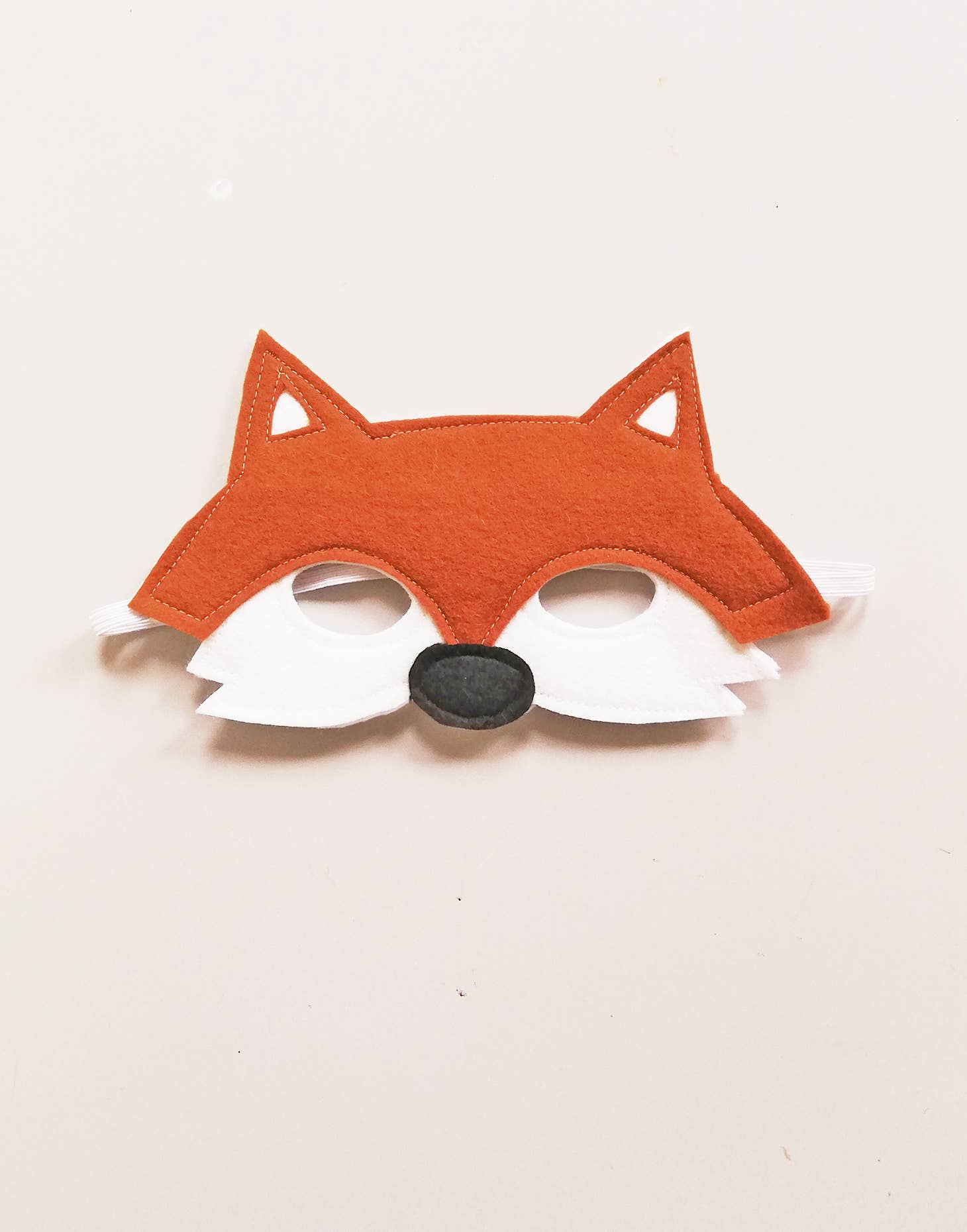 Fox mask with orange face, white whiskers, and black nose on a light beige background