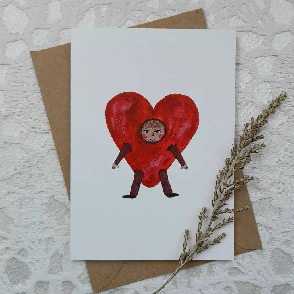 Card with a heart-shaped character on a textured surface with a brown envelope and dried plant.