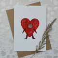 Card with a heart-shaped character on a textured surface with a brown envelope and dried plant.