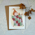 Card with illustration of a child in a quilt, surrounded by dried flowers on a textured surface.
