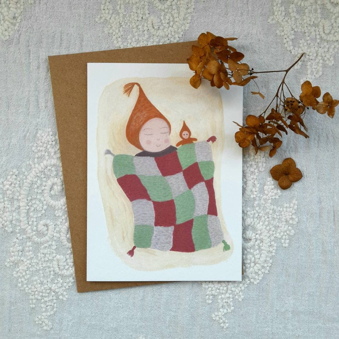 Card with illustration of a child in a quilt, surrounded by dried flowers on a textured surface.