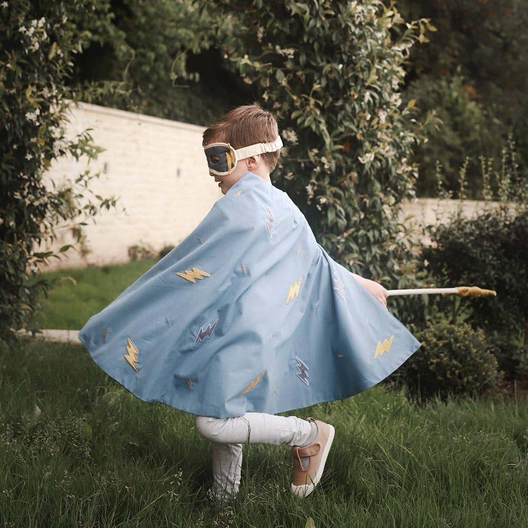 Child in a blue cape with superhero designs standing outdoors in a grassy area.