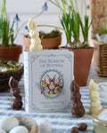 Chocolates inside a book titled 'The Burrow of Bunnies' with chocolate bunny figurines on a table with plants in the background.
