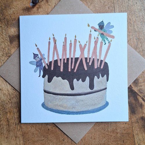 Greeting card with a birthday cake illustration featuring candles and fairies on a wooden surface.