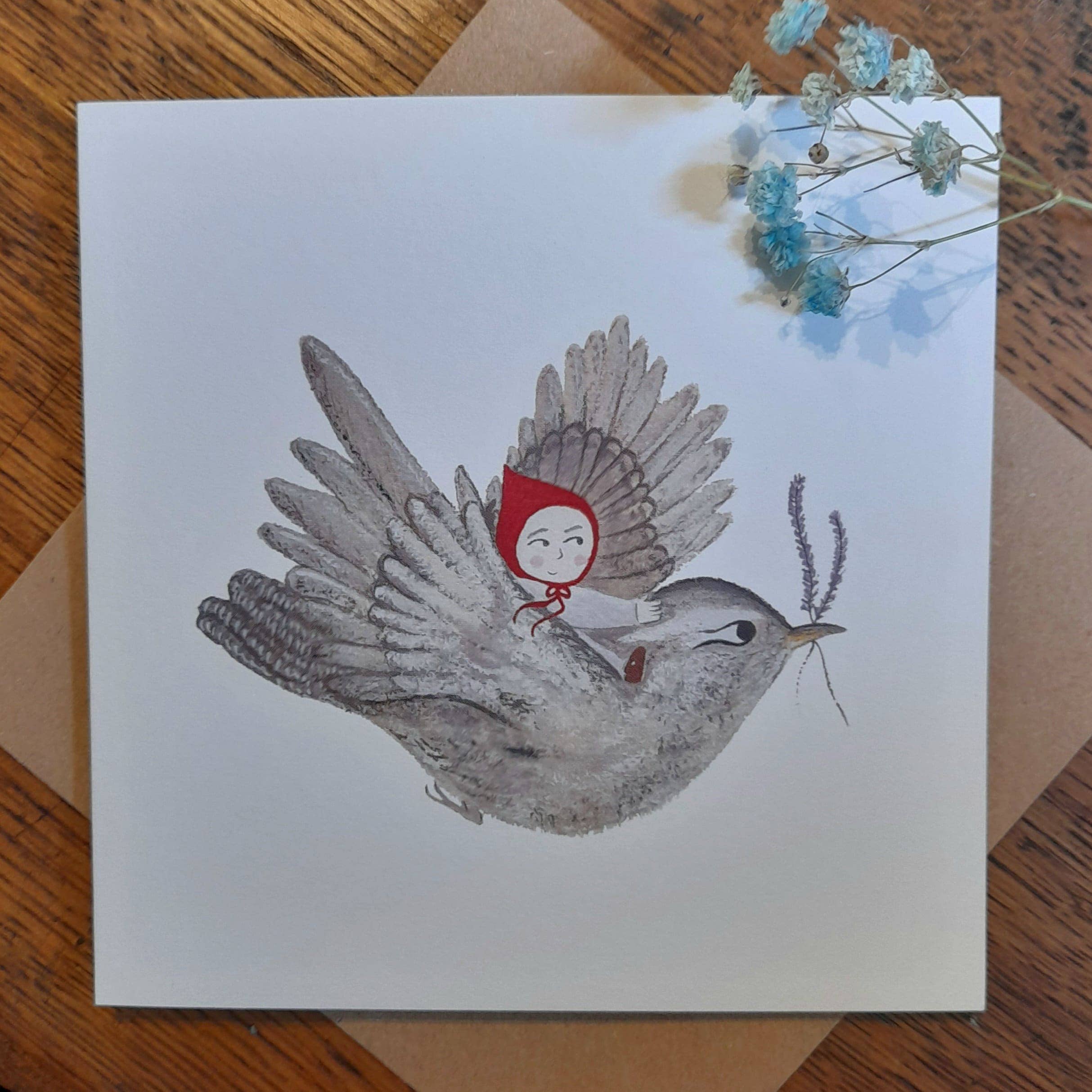 Card with illustration of a bird carrying a person in a red hood, on a wooden surface.