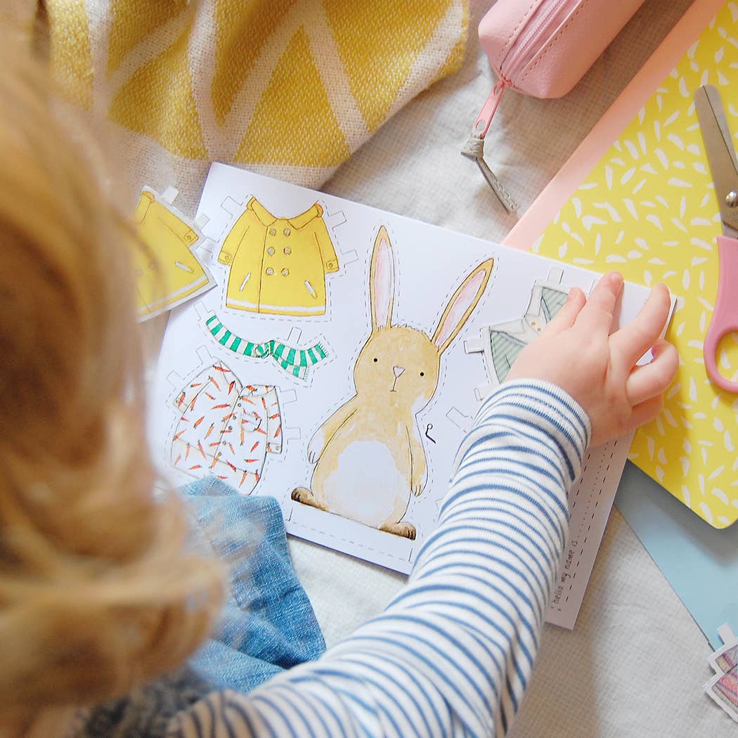 Child playing with paper dress-up clothes and a rabbit illustration on a table.
