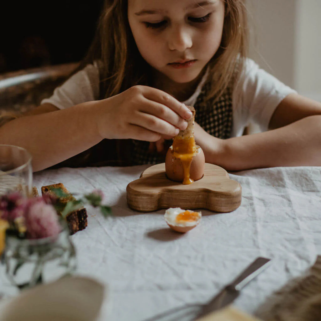 Child at a table with a wooden egg holder and a cracked egg on a white cloth.